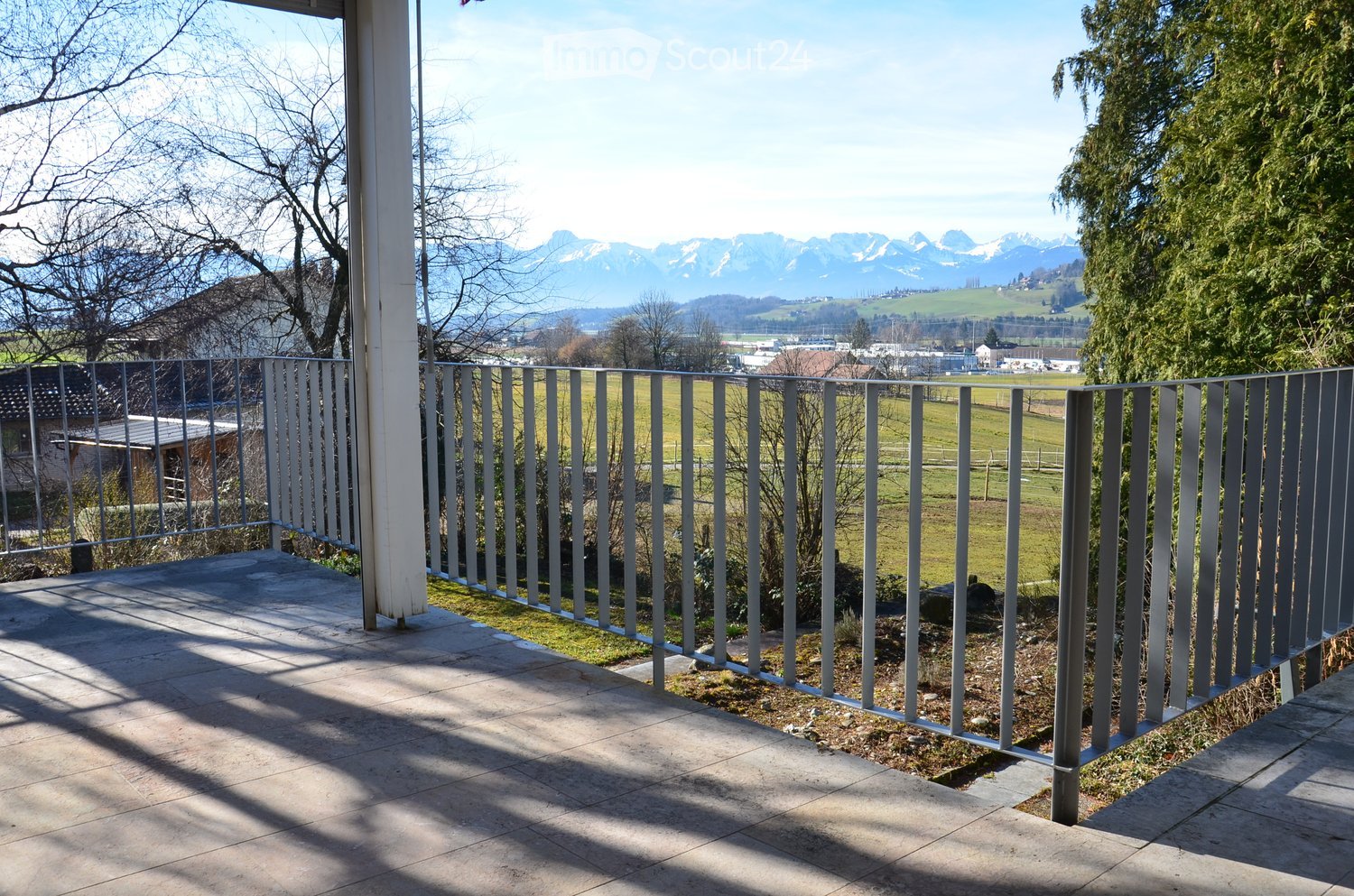 Balcony with metal railing, tiled floor, overlooking a green field and distant mountains.