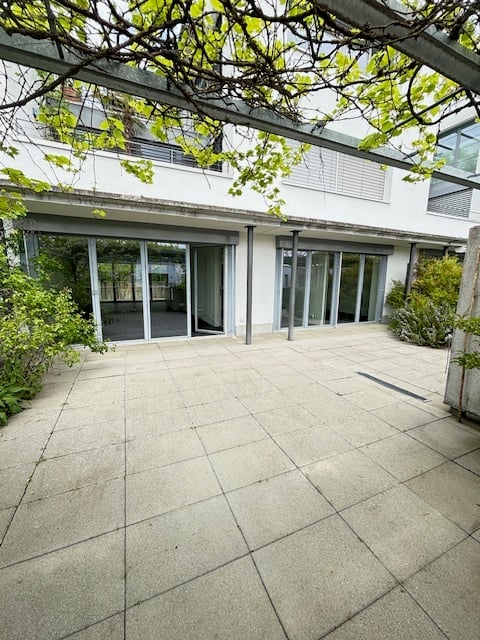A modern outdoor patio area with large glass sliding doors leading to a white building, surrounded by greenery and trees.