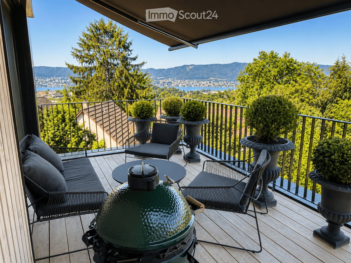 Balcony with wooden decking, black metal railing, and scenic mountain and lake view. Features a green grill, sofa, chairs, and potted plants.
