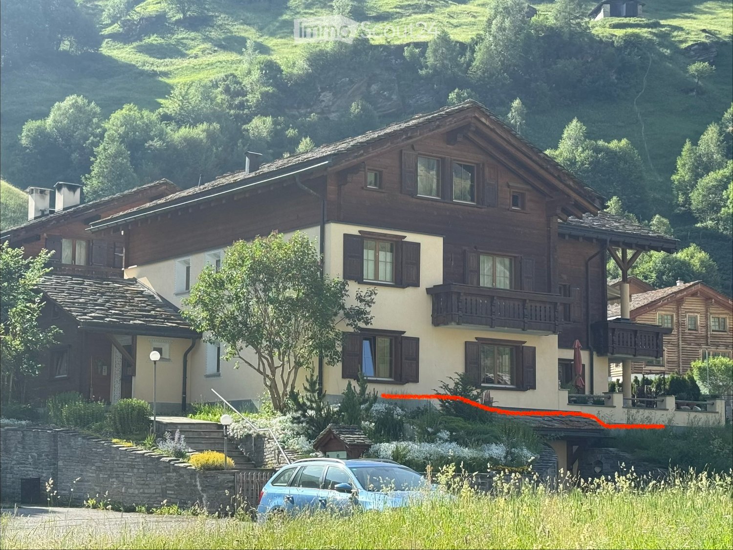 A two-story wooden house with a sloped roof, multiple windows, and balconies, surrounded by greenery and a stone wall.