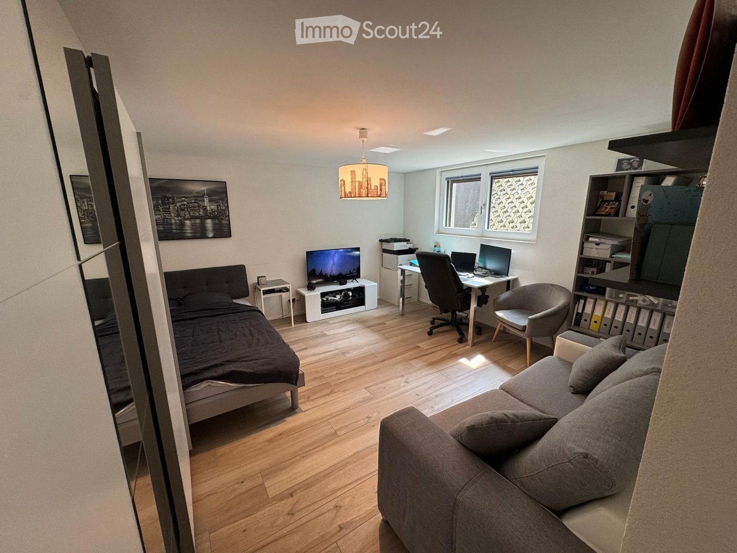 A modern, multi-functional room with a bed, sofa, desk, and shelving unit. The room features wooden flooring, large windows, and contemporary decor.