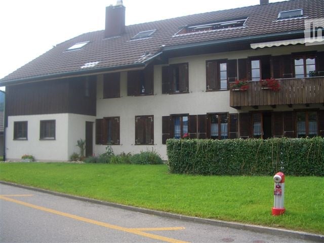 Two-story house with a red-tiled roof, white walls, and multiple windows with shutters. Features a well-maintained lawn and a small garden area.