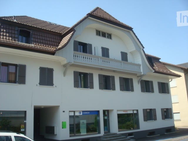 Building. A two-story white building with a brown tiled roof, multiple windows with shutters, and a small balcony on the upper floor. The ground floor features a glass entrance with a sign that reads "ZURICH".