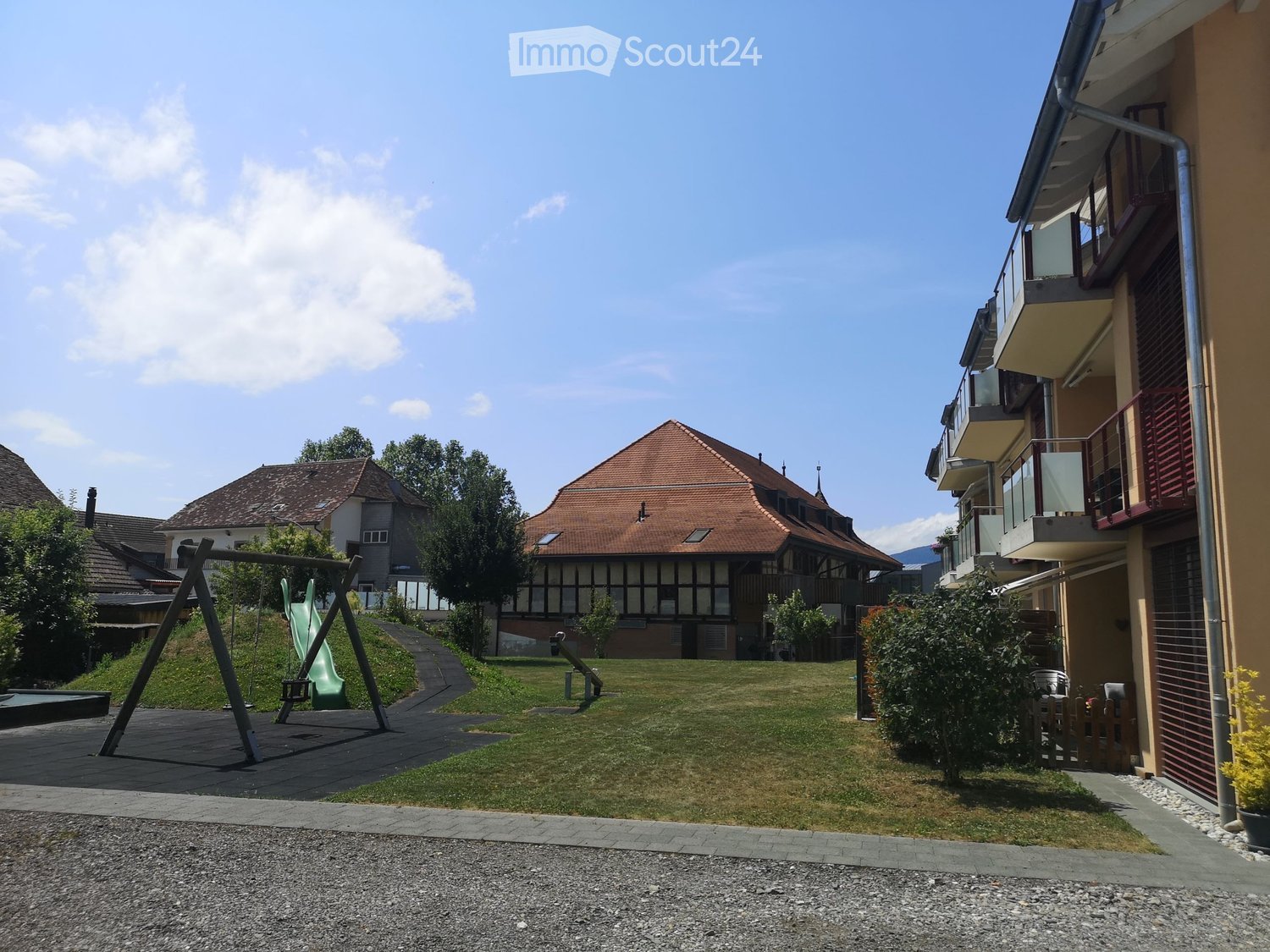 Residential area with a grassy lawn, playground equipment, and several buildings with balconies and red-tiled roofs.