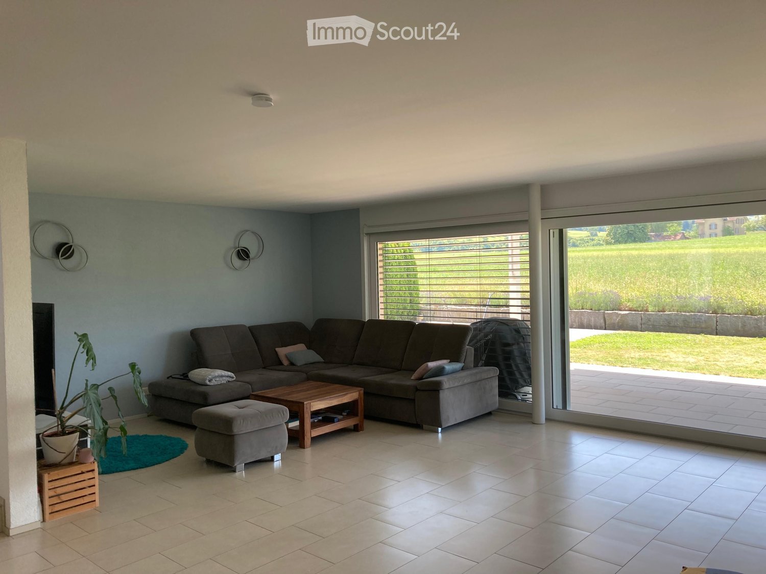 Modern living room with a grey sectional sofa, wooden coffee table, ottoman, potted plant, and wall-mounted speakers. Large windows with blinds offer a view of a green outdoor area.