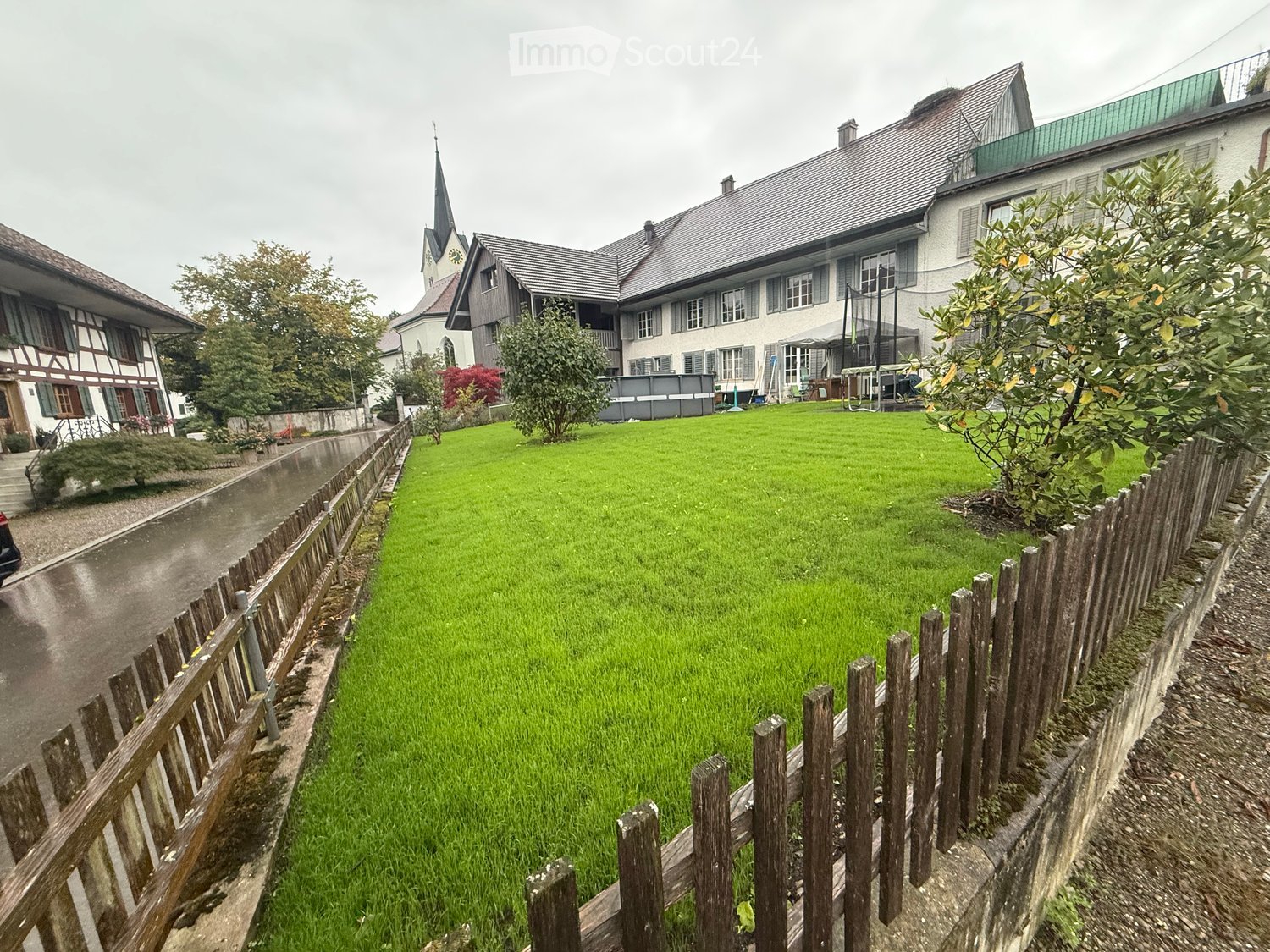 A well-maintained lawn enclosed by wooden fences, with a small stream running alongside, and several buildings and trees in the background.