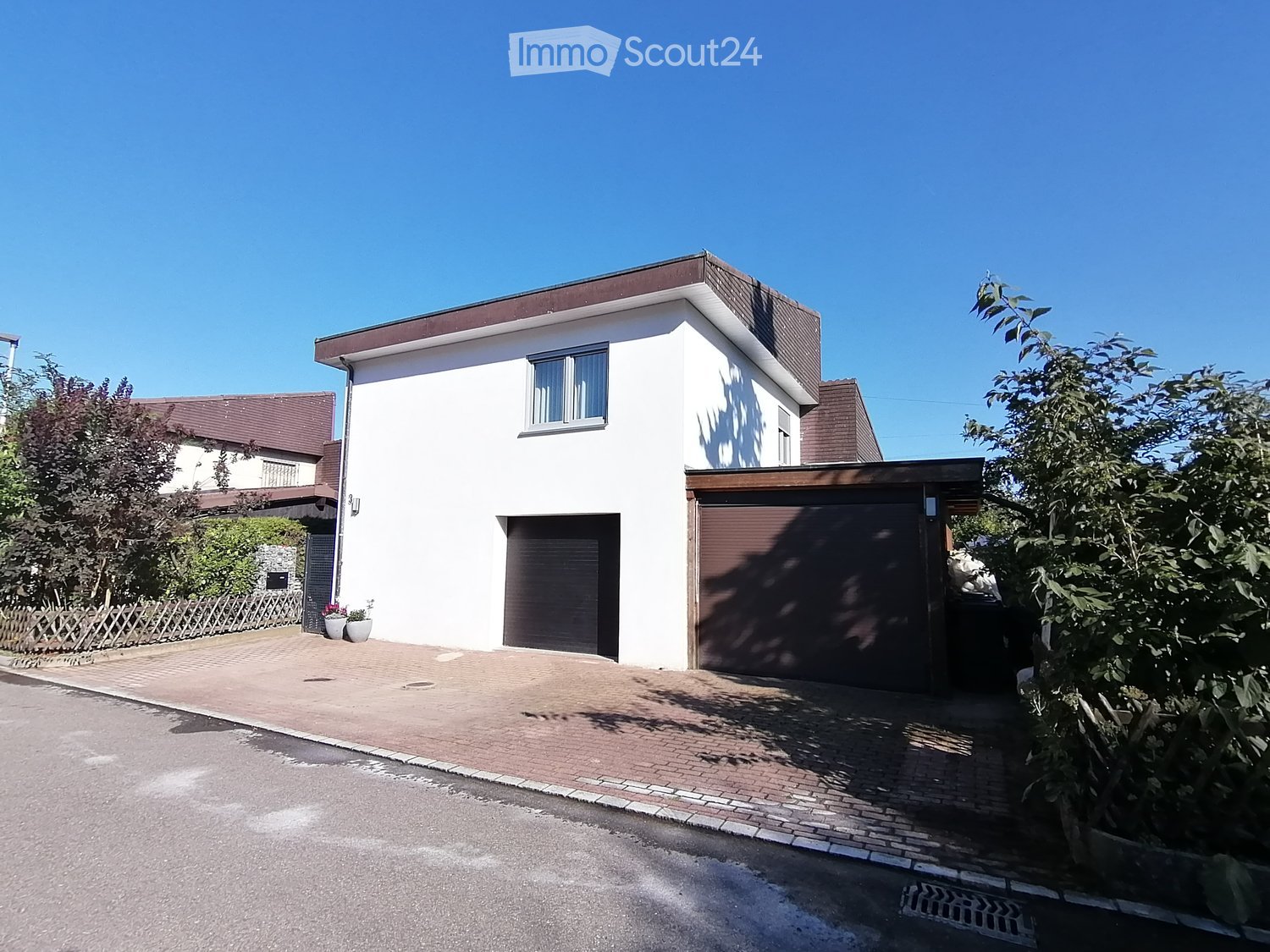 white house, brown roof, 2 garages, brick driveway, plants, trees