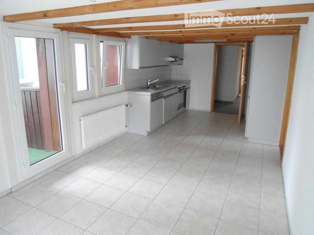 Bright, modern kitchen with white tiled flooring, white cabinets, and stainless steel appliances including a sink, faucet, oven, and dishwasher. A radiator is visible on the wall, and the space has large windows allowing natural light.