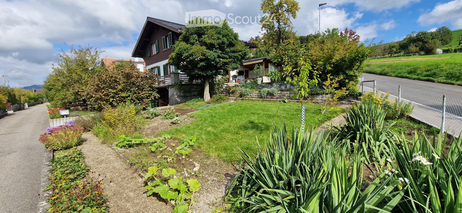 A well-maintained garden area with a variety of plants, shrubs, and flowers, leading up to a traditional Swiss house with a sloped roof and green shutters.