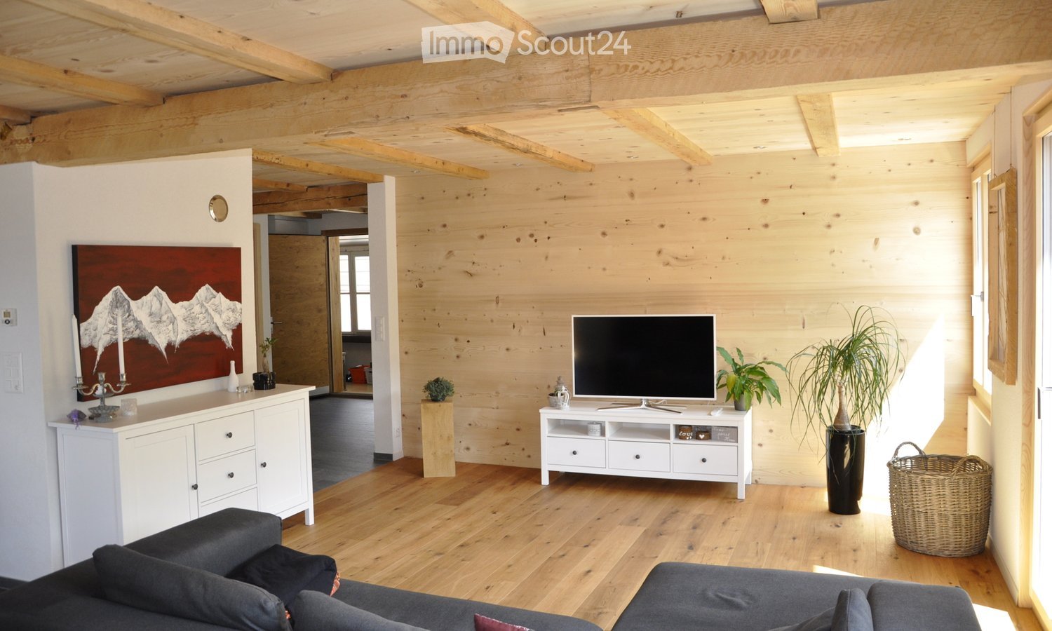 Modern living room with exposed wooden beams, light wooden walls, hardwood floor, grey sectional sofa, flat-screen TV on a white stand, decorative cabinet, potted plants, and a woven basket.