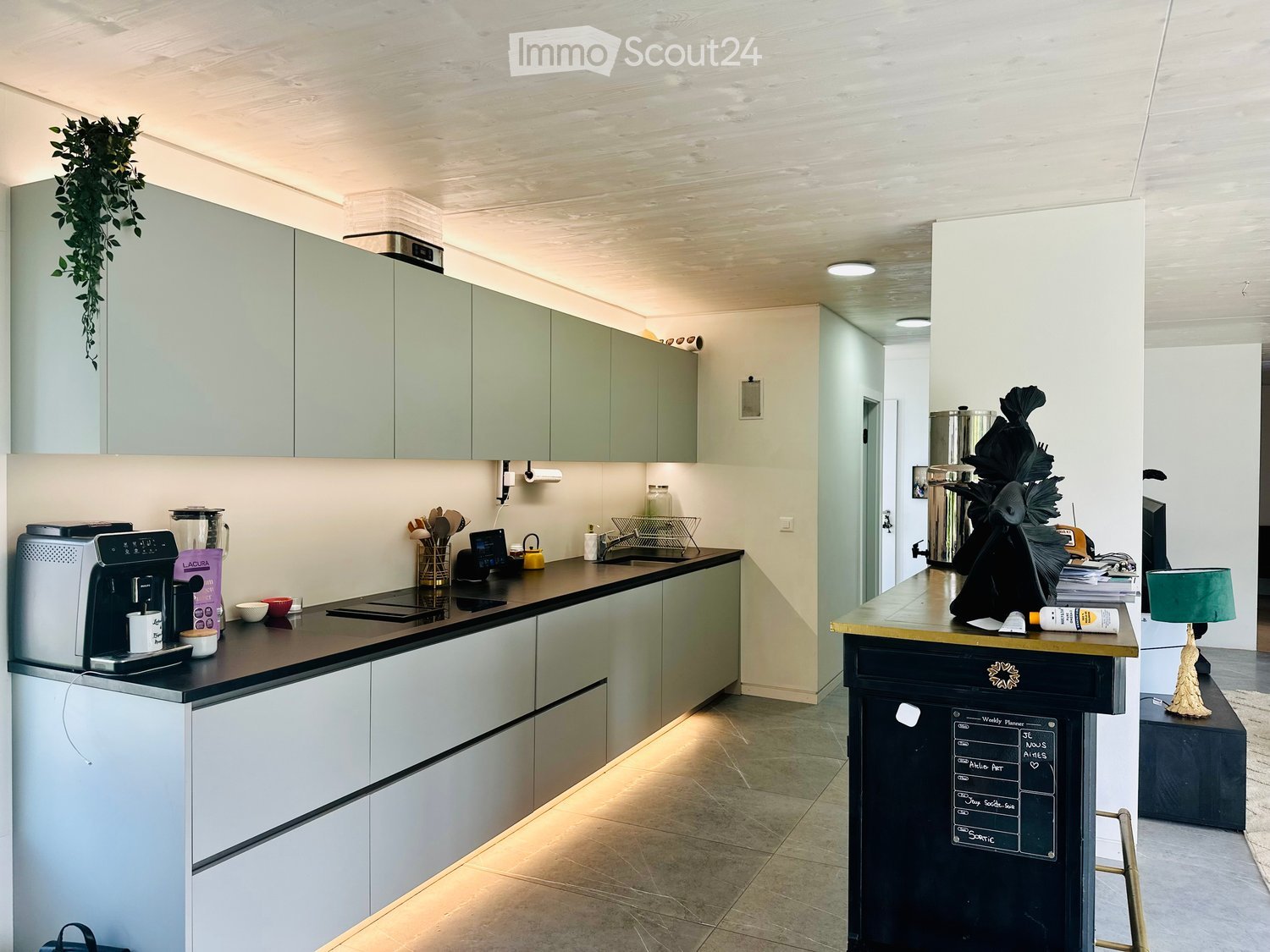Modern kitchen with light green cabinets, stainless steel appliances, and a central island with a chalkboard. Includes a coffee machine, blender, sink, and decorative items.