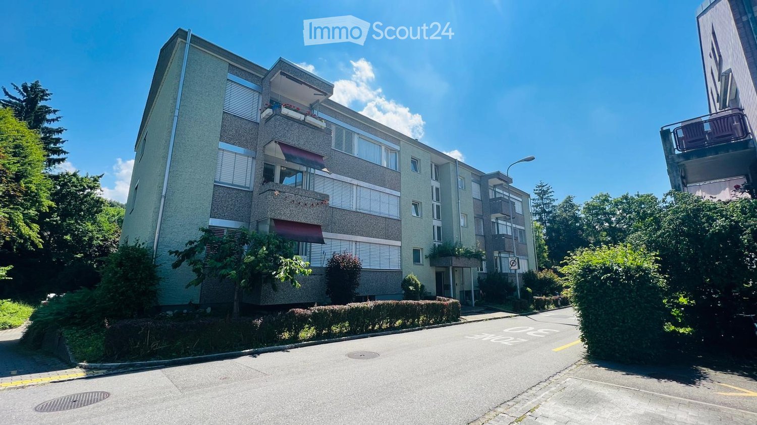 Multi-story residential building with balconies, surrounded by greenery and a clear blue sky.