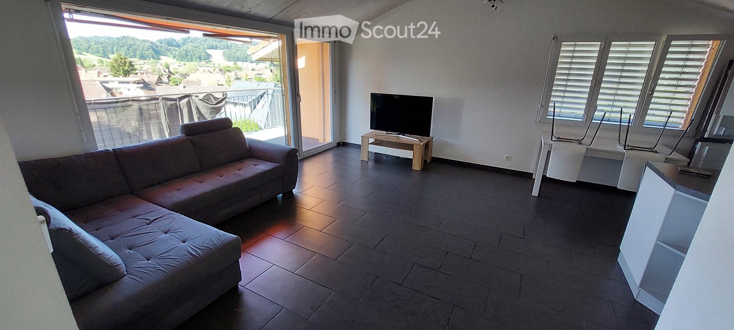 Modern living room with dark tiled flooring, a brown sectional sofa, a small wooden coffee table with a TV, and a view of the surrounding landscape through large windows and a sliding glass door leading to a balcony.