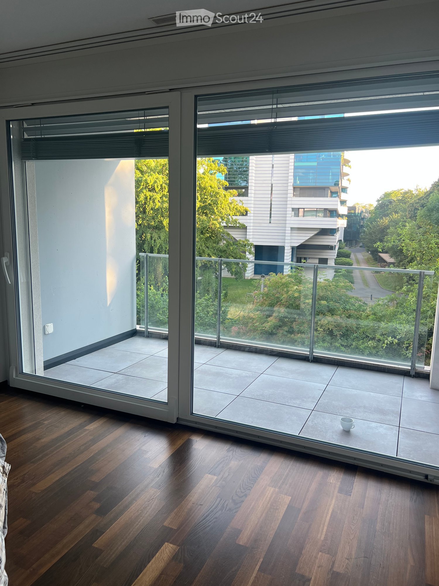 A modern, well-lit room with wooden flooring and large glass doors leading to a balcony. The balcony features a tiled floor and a glass railing, offering a view of greenery and modern buildings.