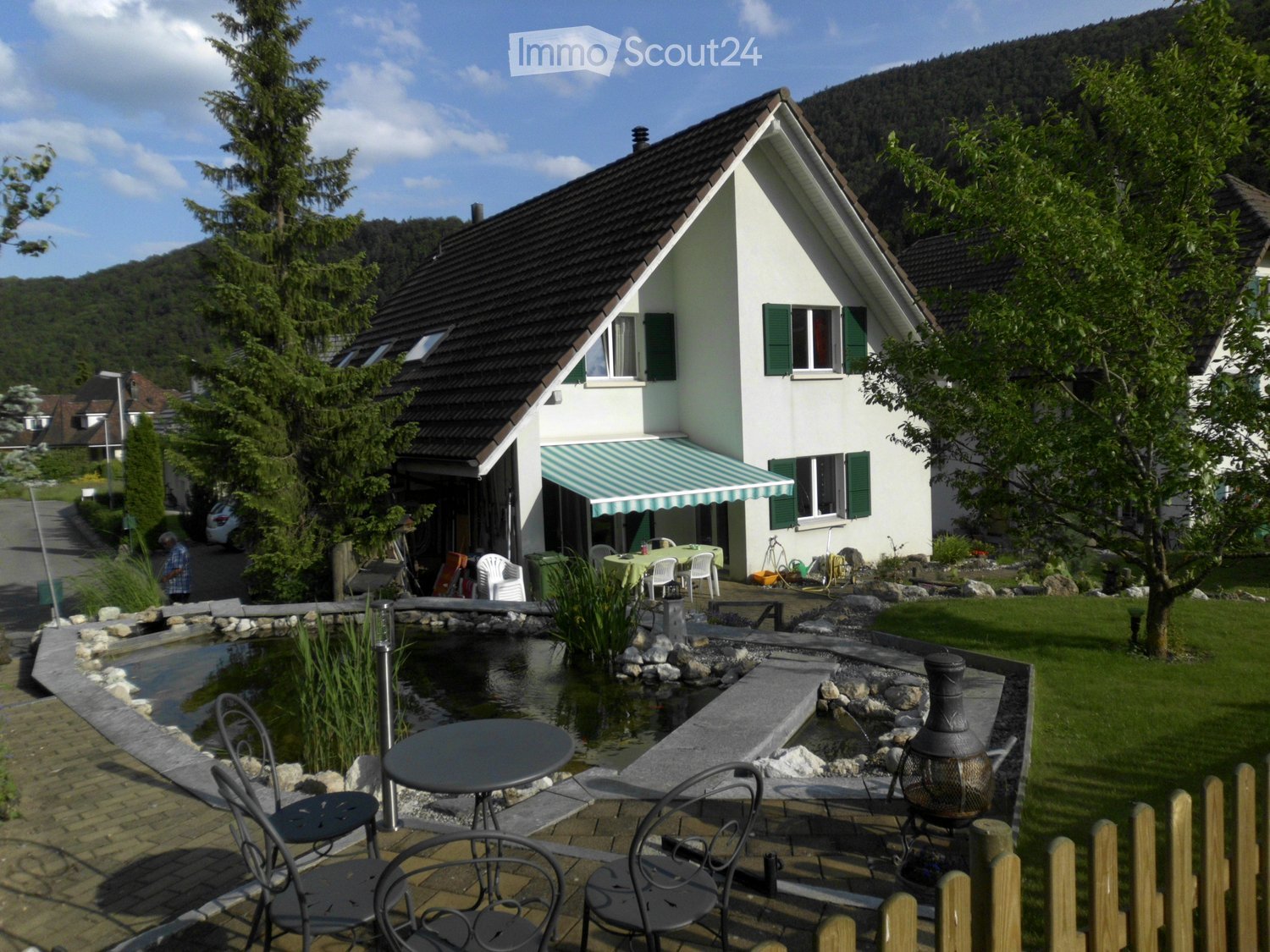 A modern two-story house with a well-maintained garden, featuring a small pond, patio furniture, and a covered outdoor seating area.