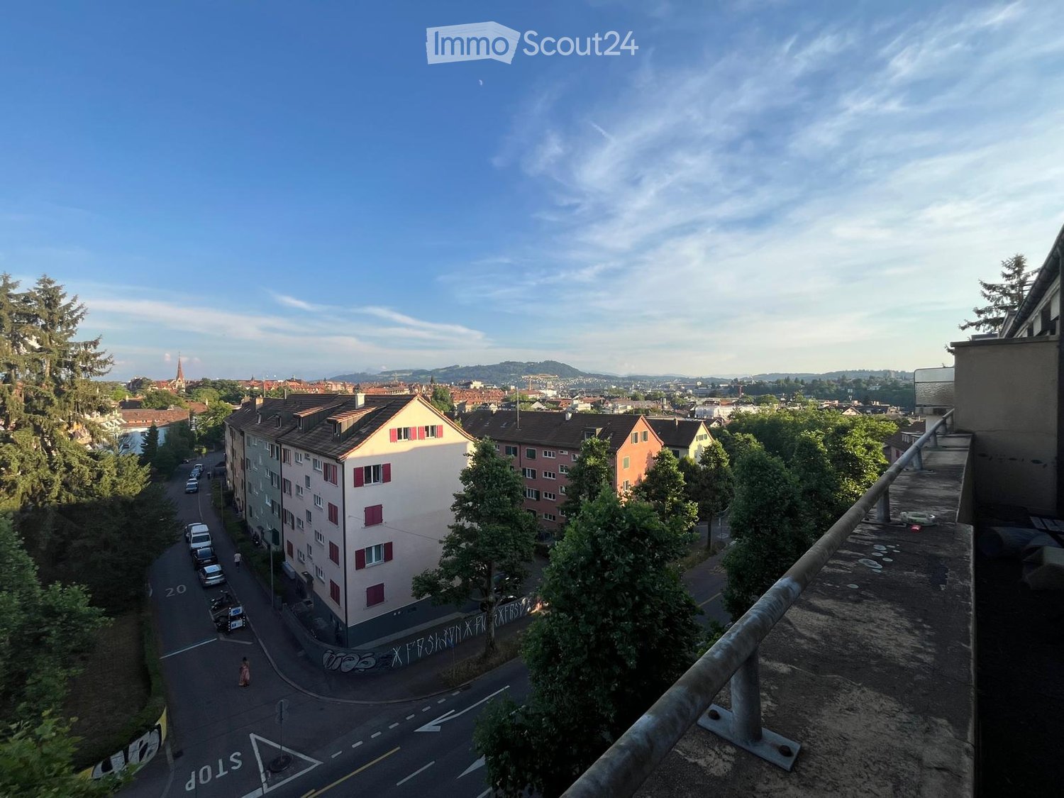 Balcony overlooking a city street with buildings, trees, and parked cars. The view includes a distant church spire and hills.