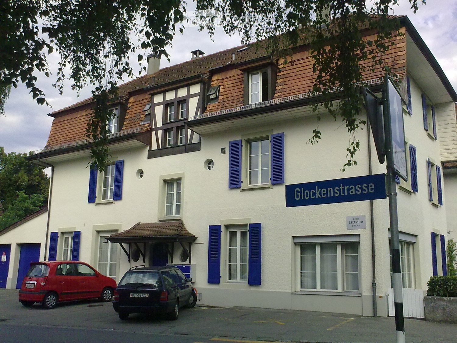White building with brown roof, blue shutters, two cars parked in front