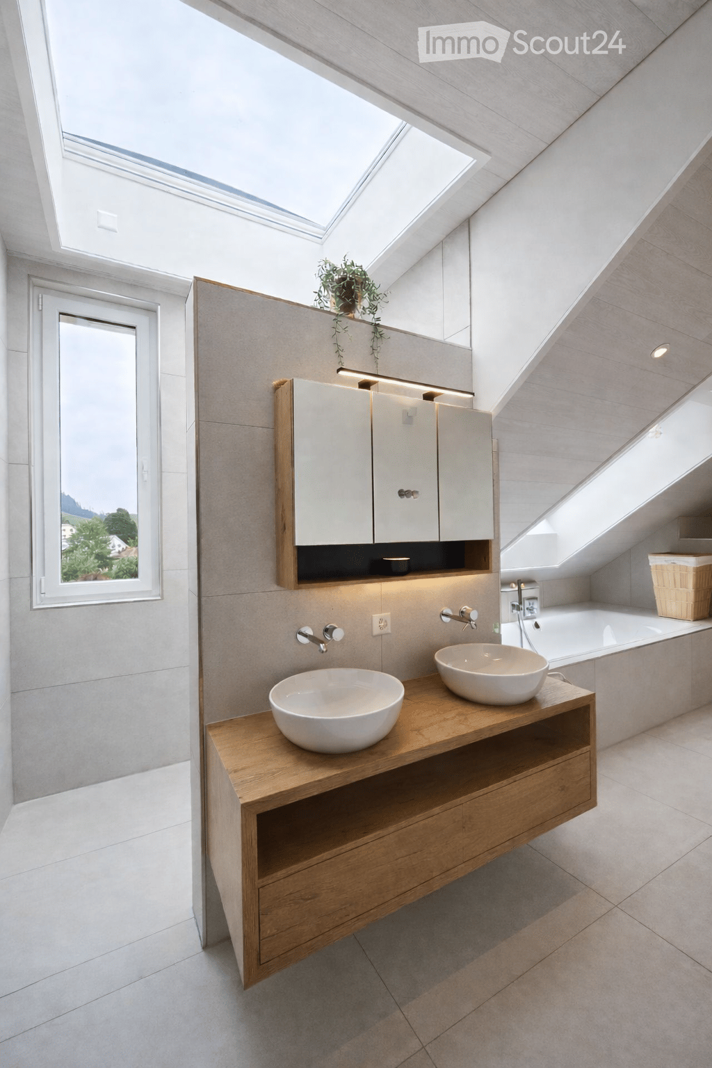 Modern bathroom with two sinks on a wooden vanity, a large mirror with built-in cabinets, a bathtub, and a skylight window providing natural light.