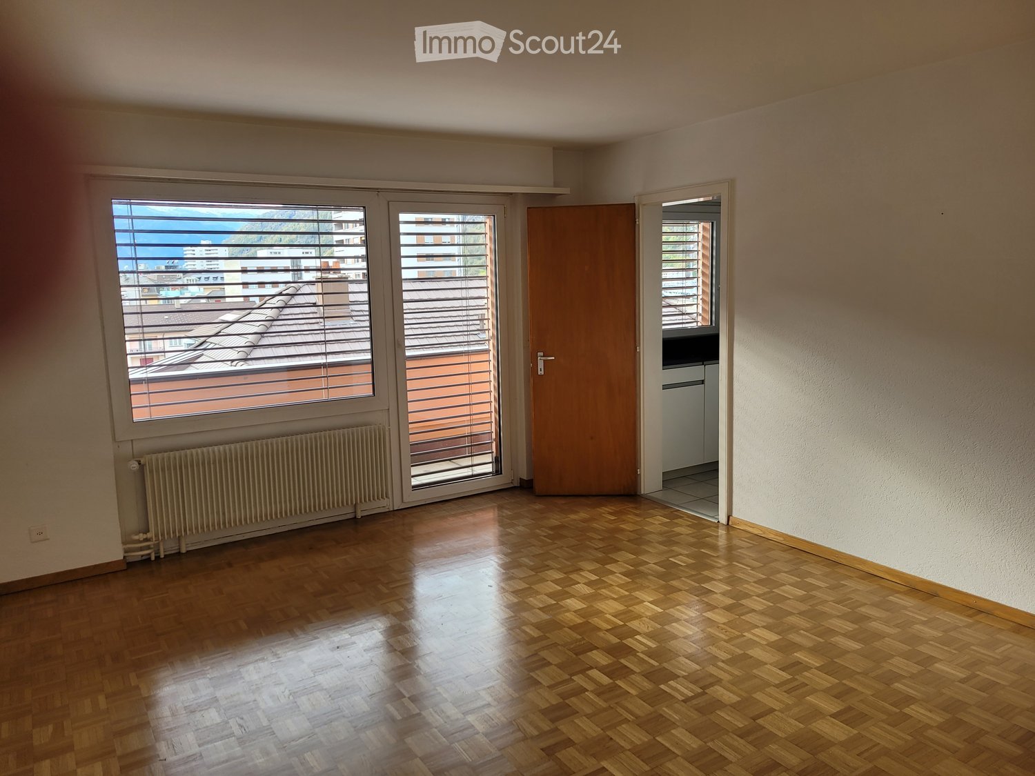 Empty room with wooden flooring, large windows with blinds, radiator, and a door leading to another space.