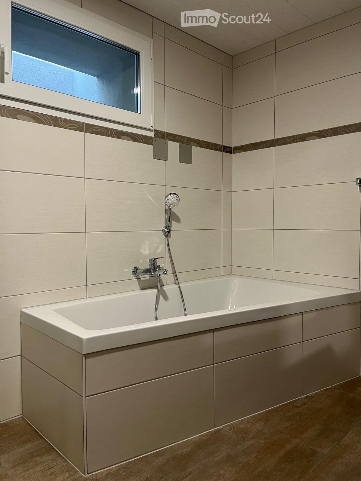 Modern bathroom with a bathtub, faucet, and showerhead. Light-colored tiles and a window provide natural light.