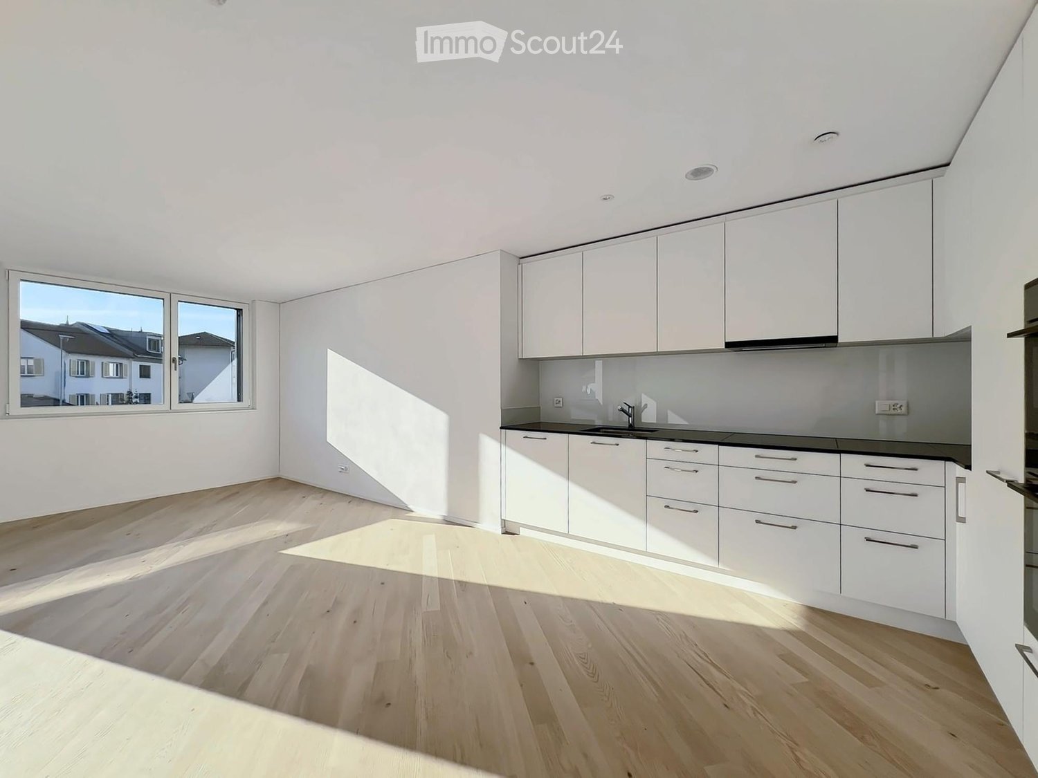 Modern kitchen with white cabinets, light wooden flooring, and a large window offering a view of the street.