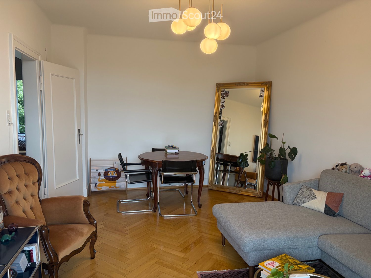 Living room with wooden flooring, a grey sofa, a brown leather armchair, a round wooden table with chairs, a large ornate mirror, and a potted plant. A hanging light fixture is above the table.