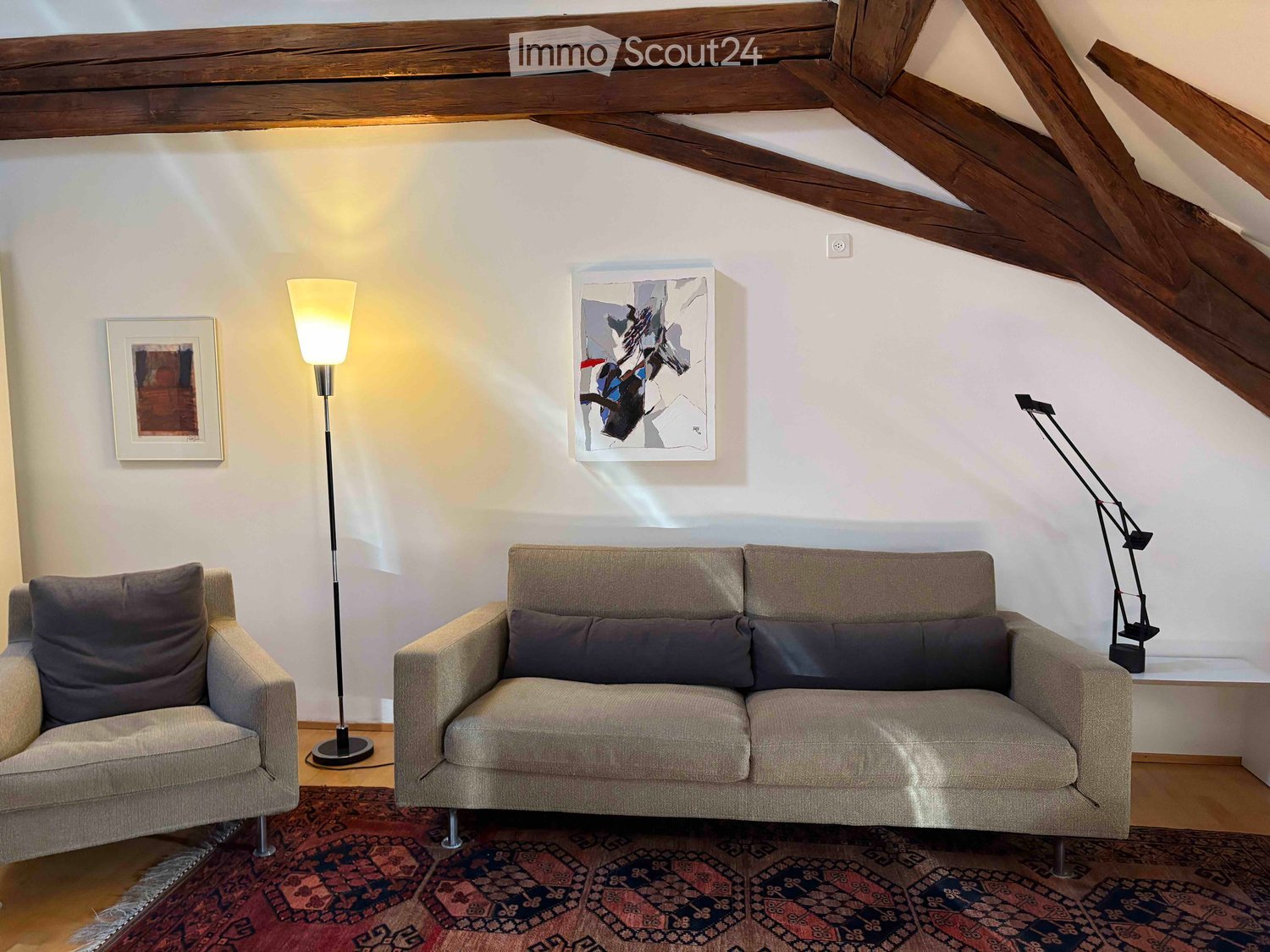 Living room with exposed wooden beams, a beige sofa, an armchair, a floor lamp, a table lamp, and two picture frames on the wall.