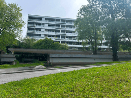 Multi-story residential building with multiple balconies and windows, surrounded by greenery and a paved pathway.