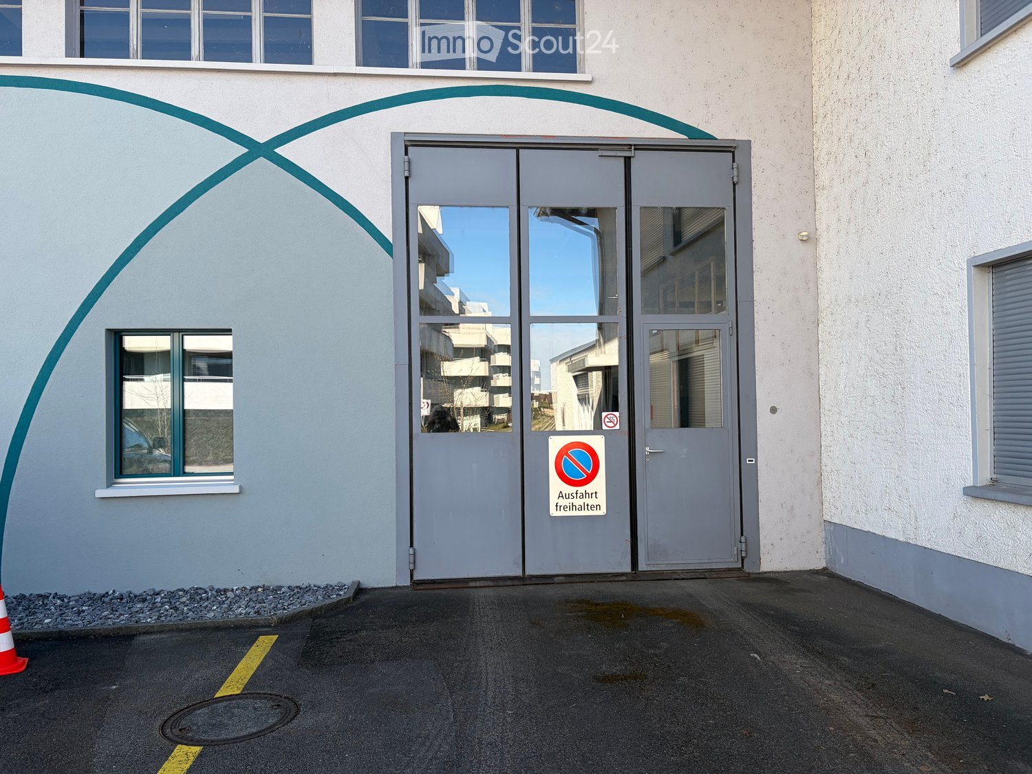 Exterior view of a building entrance with a large grey door, windows, and a no-parking sign.