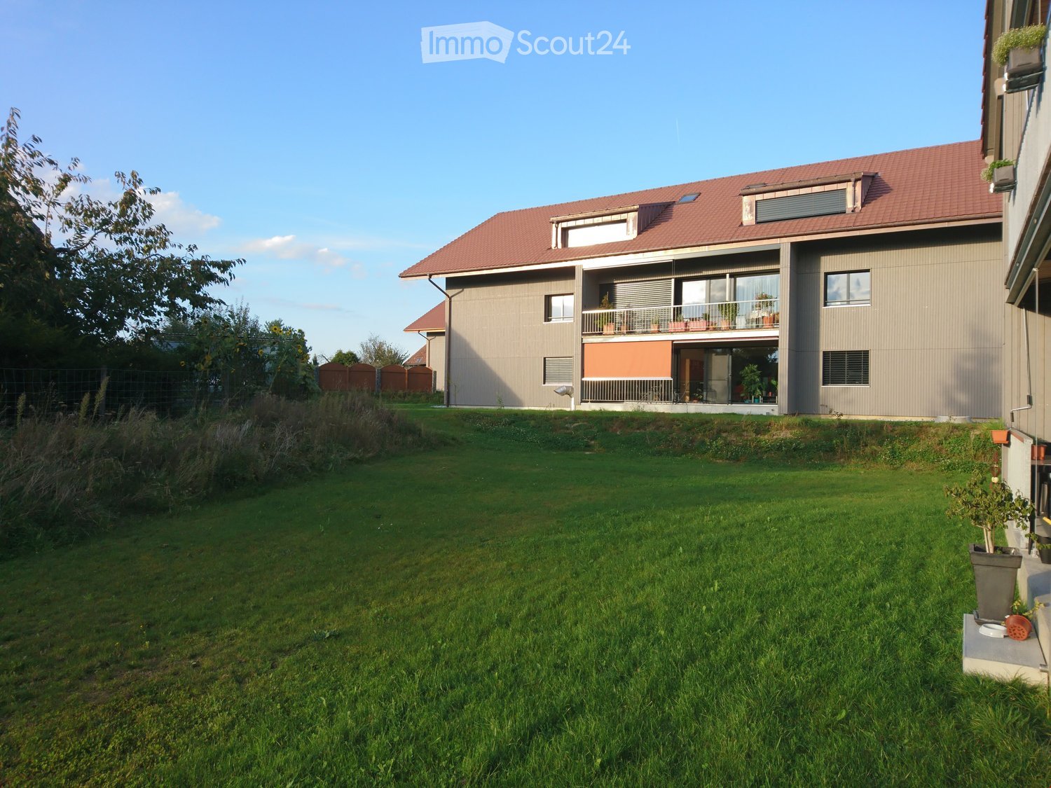 Lush green lawn in front of a modern two-story house with a red-tiled roof, large windows, and a balcony with outdoor furniture.