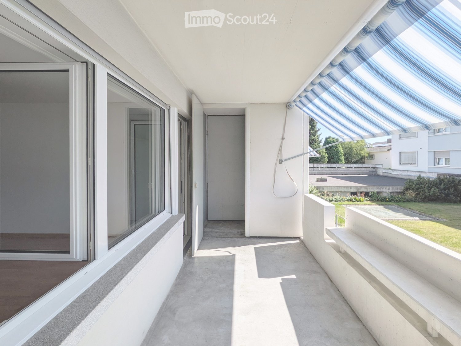 Modern balcony with large glass doors, white walls, and a striped awning. The space is clean and open, with a view of a green lawn and neighboring buildings.