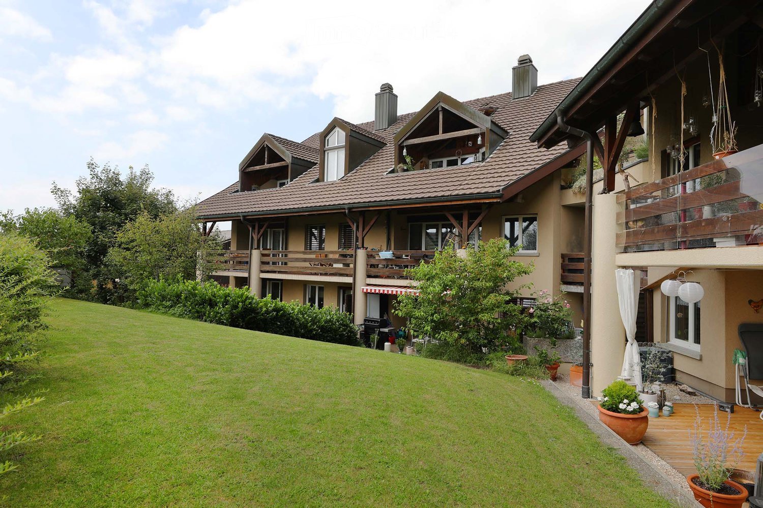 A two-story house with a tiled roof, multiple balconies, and a well-maintained garden with potted plants and a wooden deck.