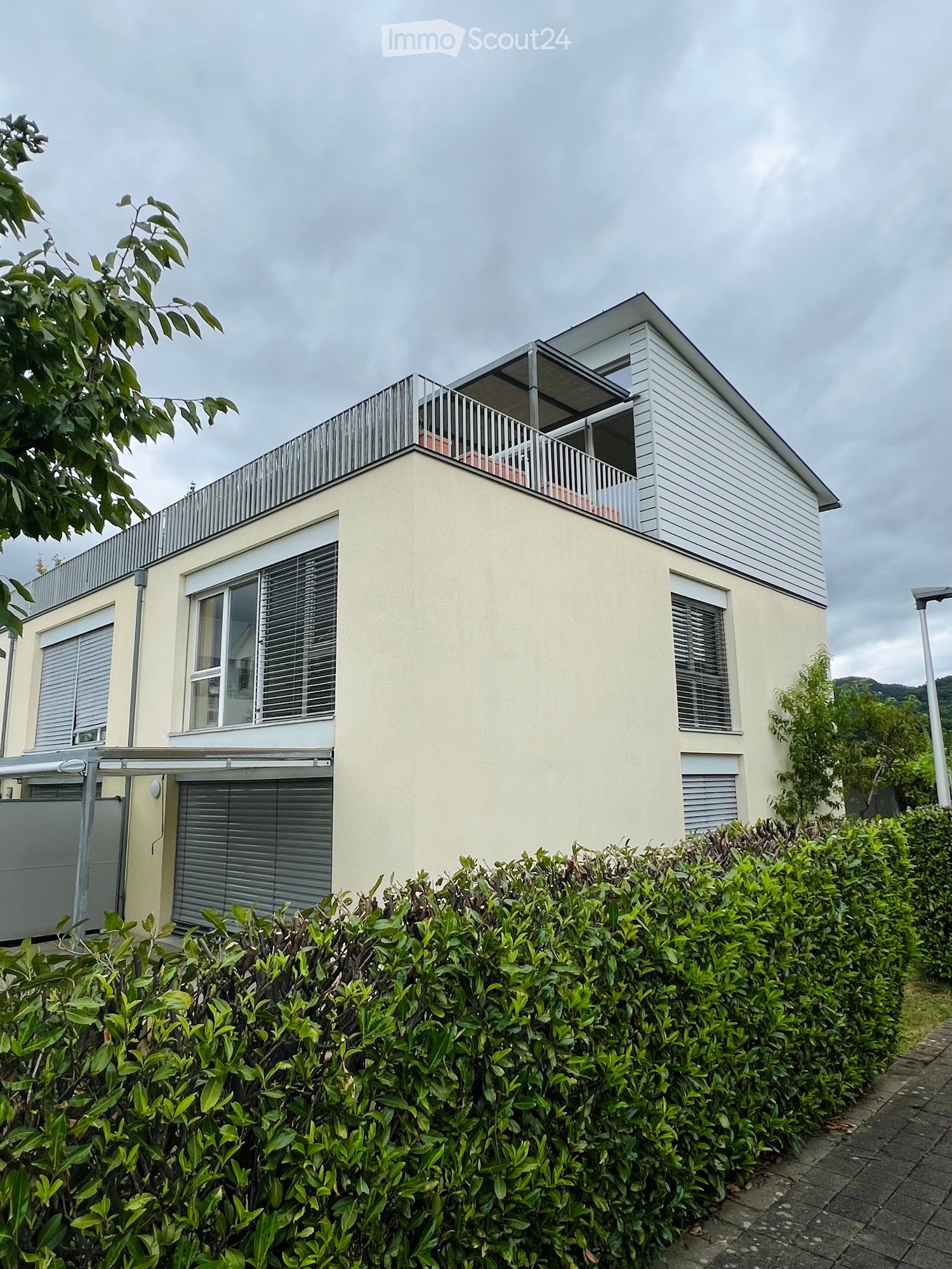 Two-story house with a modern design, featuring a balcony on the upper level, large windows, and a well-maintained hedge in the front yard.