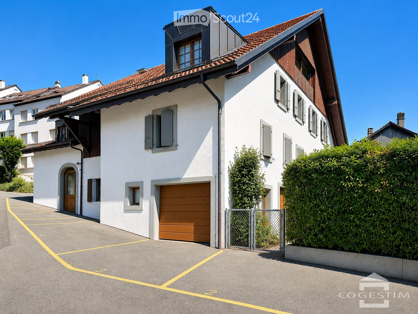 Two-story house with white exterior, wooden garage door, multiple windows, and a fenced garden area.