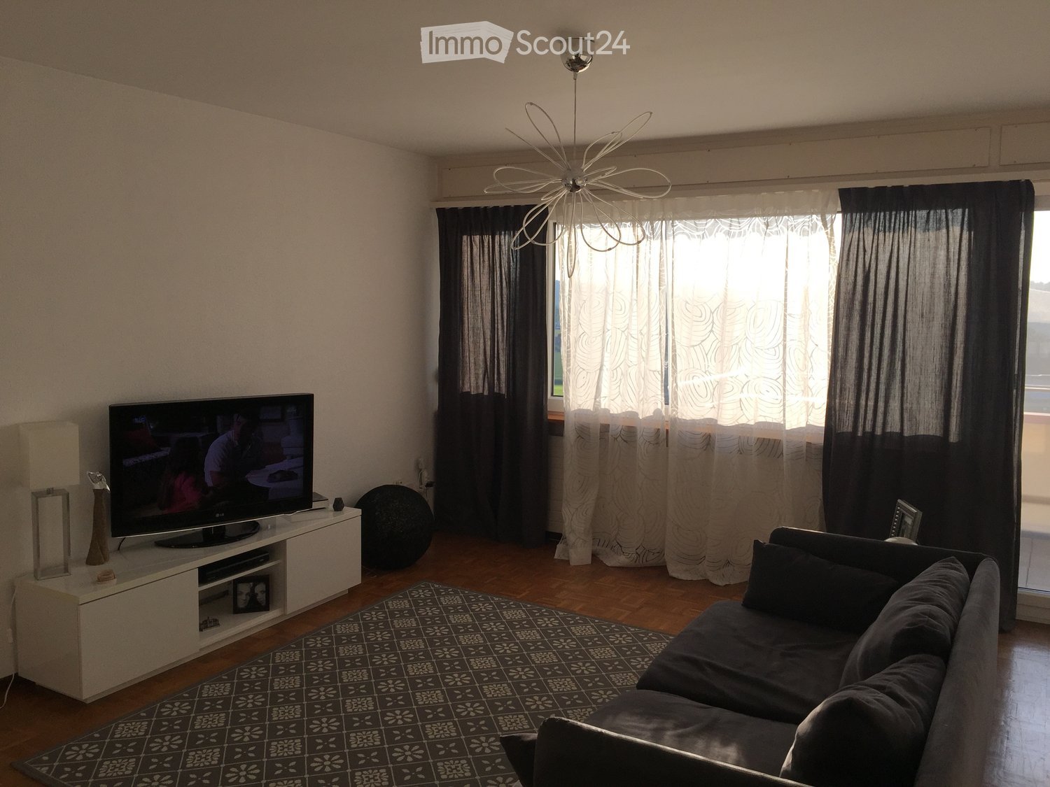 Modern living room with a grey sectional sofa, a flat-screen TV on a white stand, a patterned rug, and large windows with dark curtains. A decorative lamp hangs from the ceiling.