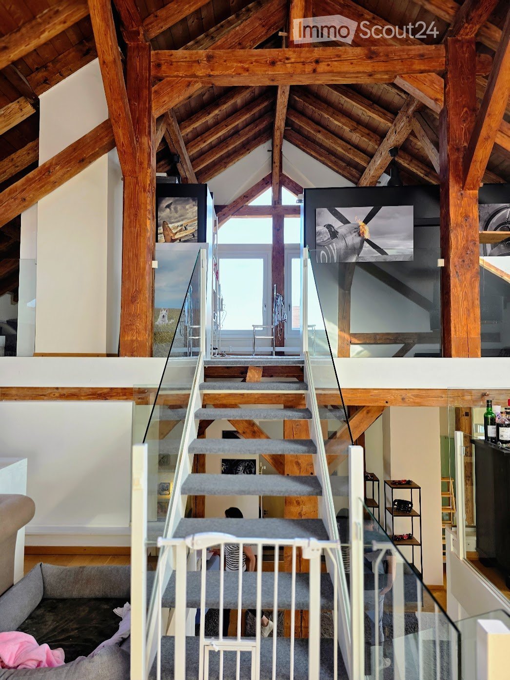 Open-plan living area with exposed wooden beams, a modern staircase with glass railings, a comfortable sofa, and a small table with chairs. The space features large windows and a high, slanted wooden ceiling.