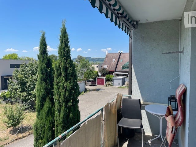 Balcony with a striped awning, a small table, and a chair, overlooking a residential area with trees and a parking lot.