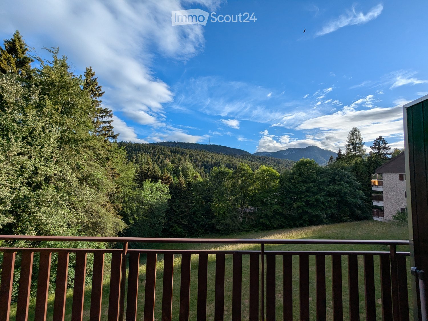 Balcony with wooden railing overlooking a lush green landscape, trees, and distant mountains under a clear blue sky.