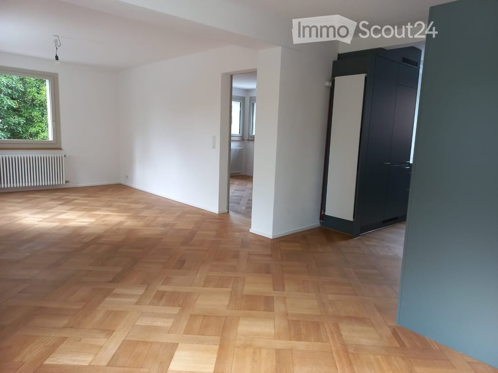Empty room with wooden parquet flooring, white walls, and large window with radiator below. No furniture or equipment present.
