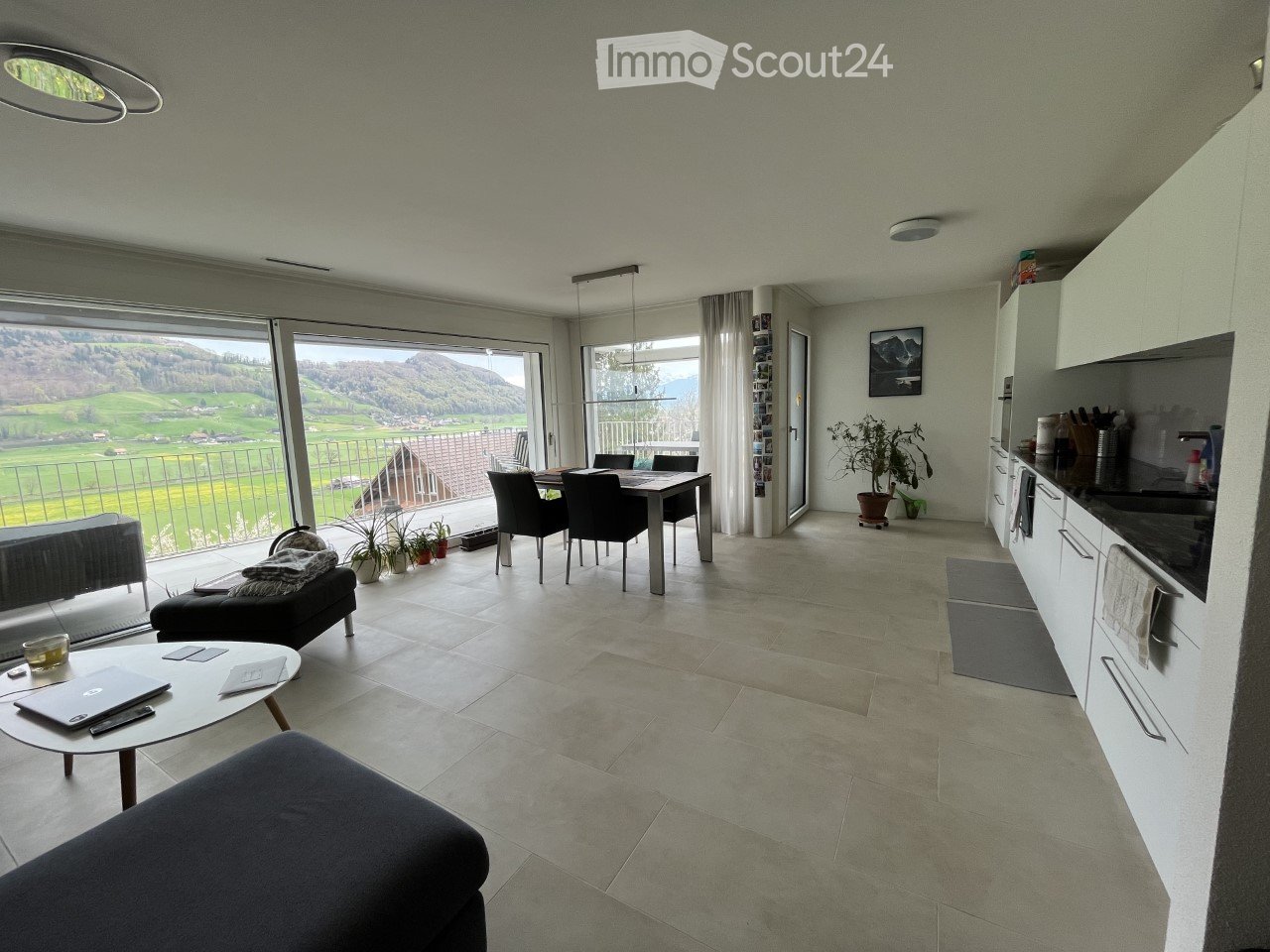 Open-plan living and dining area with large windows offering a scenic view, featuring a sofa, coffee table, dining table, chairs, and a modern kitchen area.