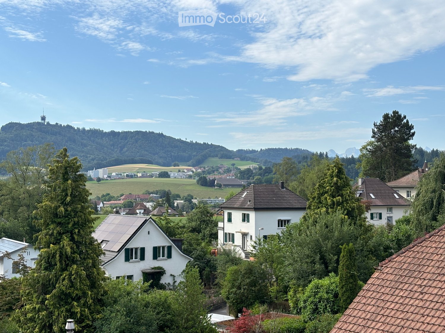 Residential houses with pitched roofs, surrounded by trees and greenery, with a scenic valley and hills in the background.