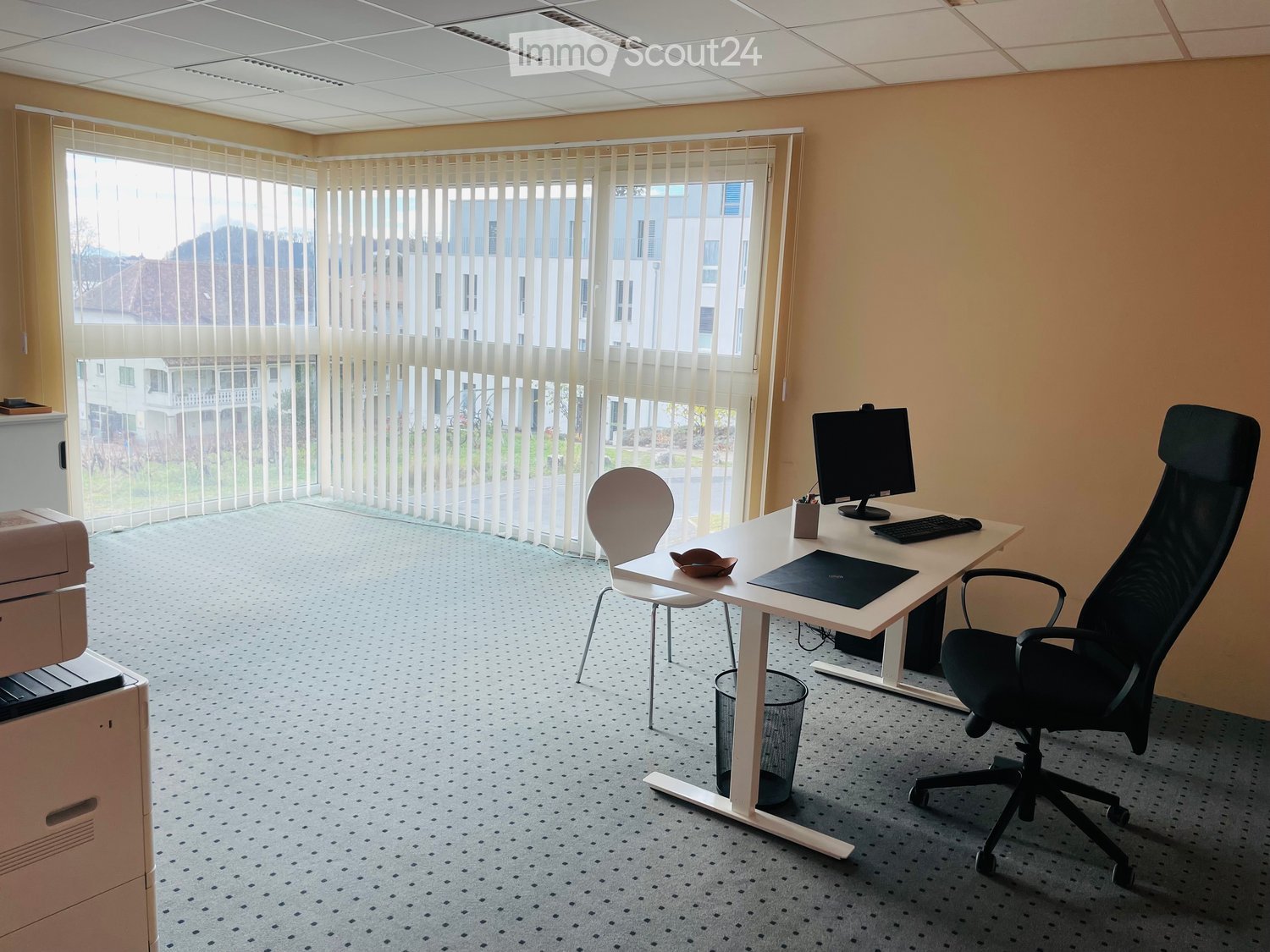 Office space with a white desk, black office chair, computer, keyboard, printer, and trash can. Large windows with vertical blinds provide natural light.