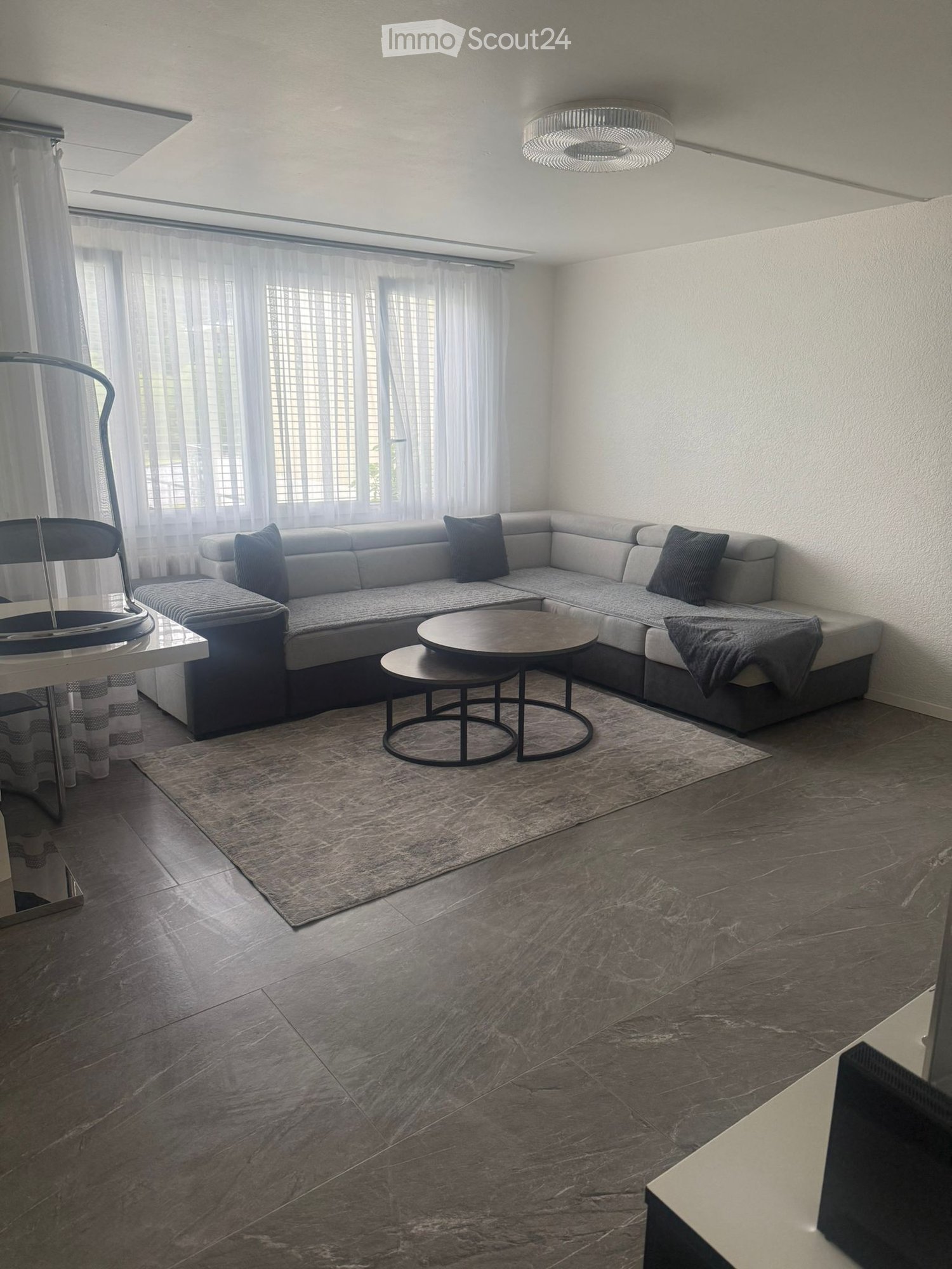 Modern living room with a grey sectional sofa, coffee table, side table, and a large window with sheer curtains. The room features a marble floor and a contemporary chandelier.