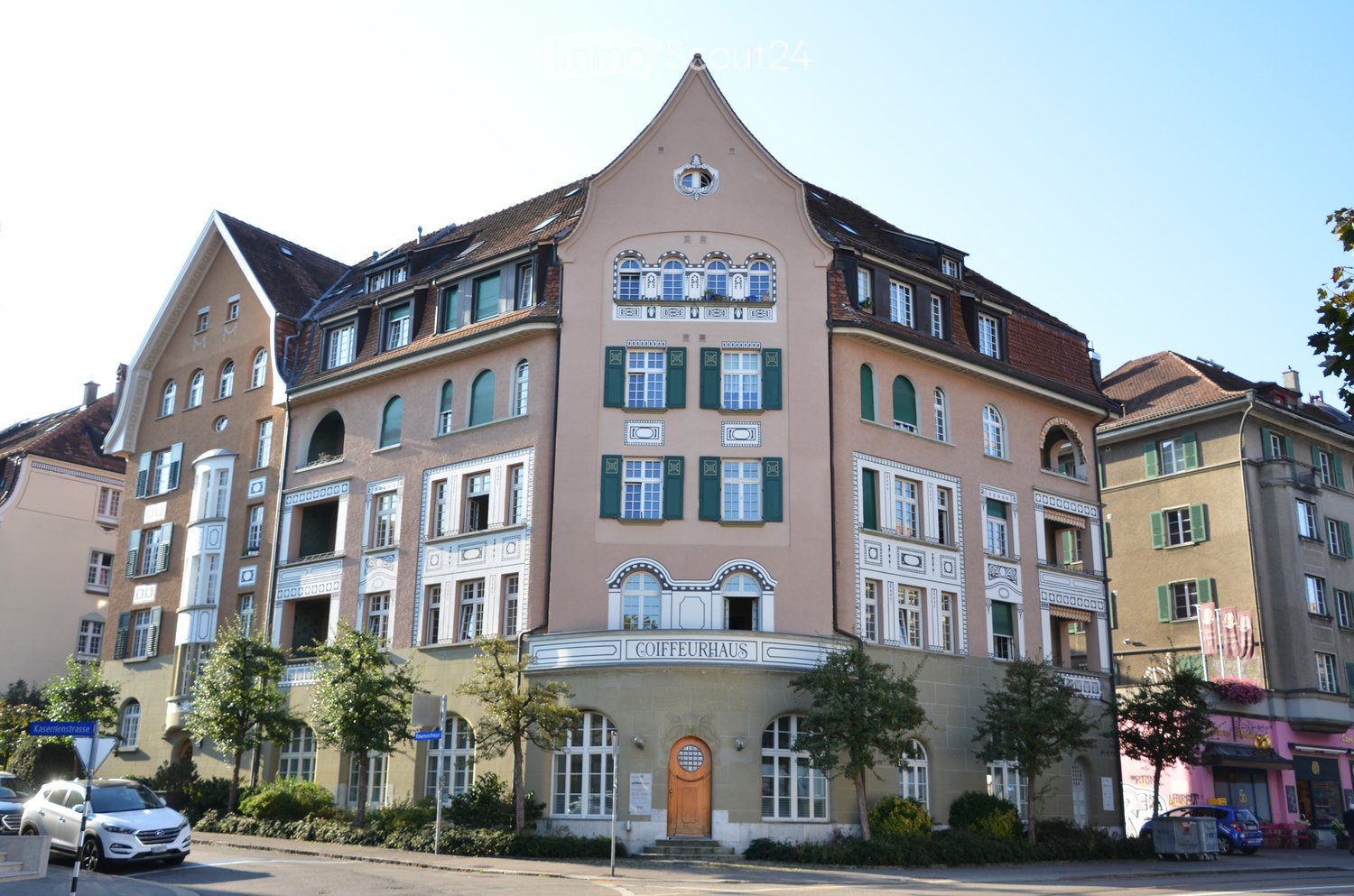 A multi-story building with ornate architectural details, including decorative window frames, green shutters, and a prominent central entrance with a sign reading "COIFFEURHAUS". The building features a mix of pastel-colored facades and is surrounded by t