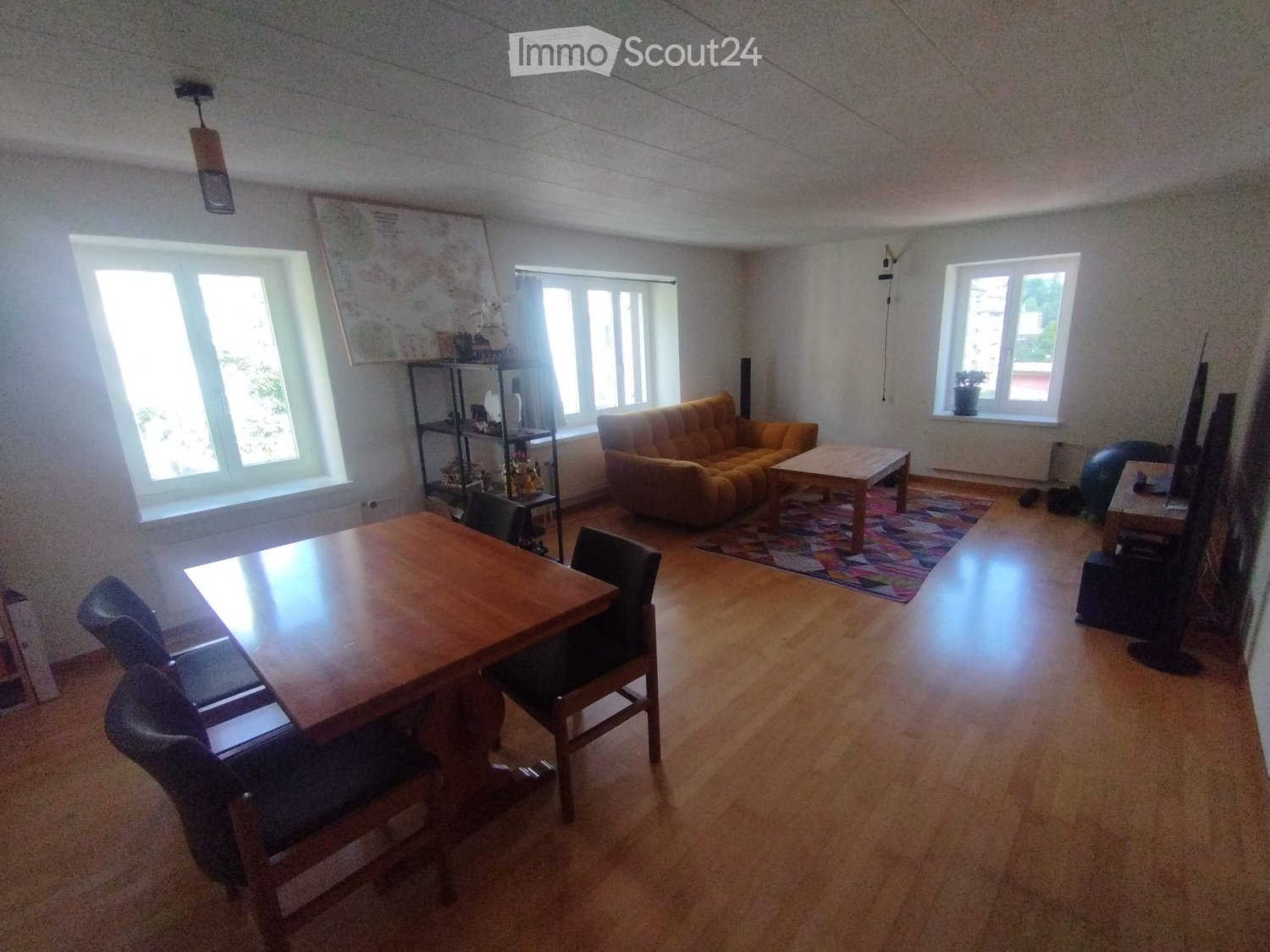 A spacious living and dining area with wooden flooring, large windows, a sofa, coffee table, dining table with chairs, and a shelf with decorative items.