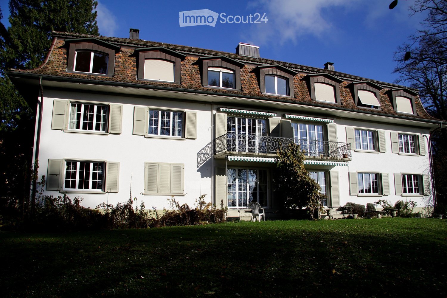 A two-story white house with a tiled roof, multiple windows with shutters, and a balcony on the upper floor. The house is surrounded by a well-maintained lawn and some outdoor furniture near the entrance.