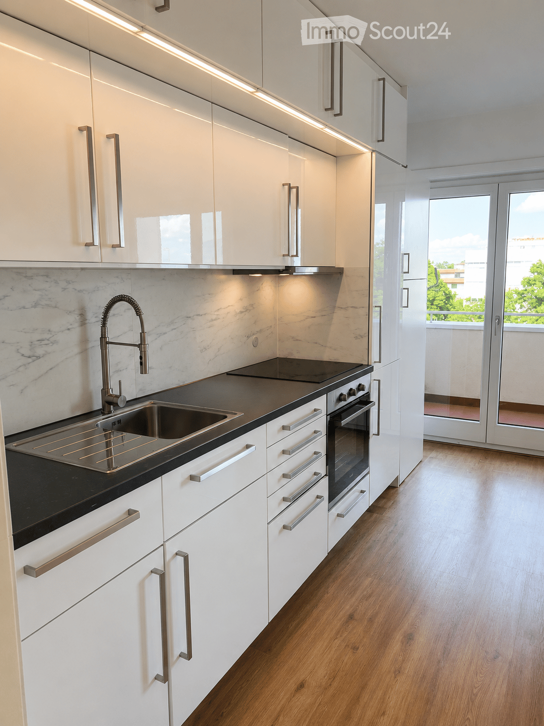 Modern kitchen with white cabinets, black countertops, a stainless steel sink, and a built-in oven. The space features wooden flooring and large windows with a view of greenery.