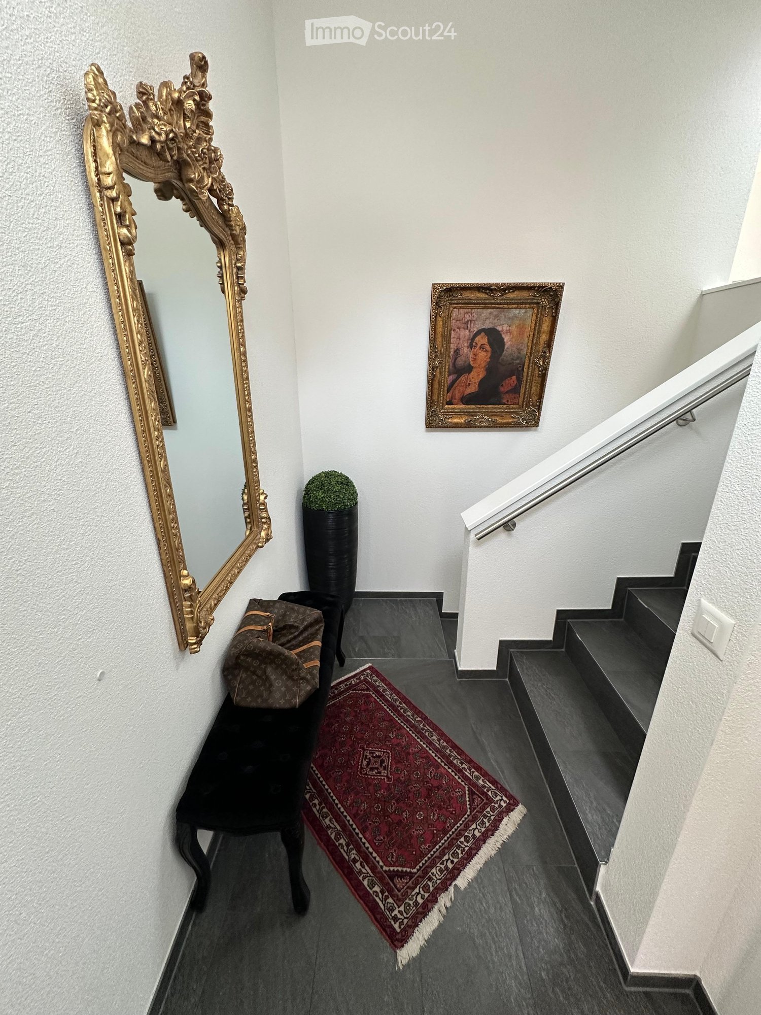 Hallway with dark tiled floor, ornate gold-framed mirror, framed painting, small potted plant, red patterned rug, and a black chair with a bag on it.