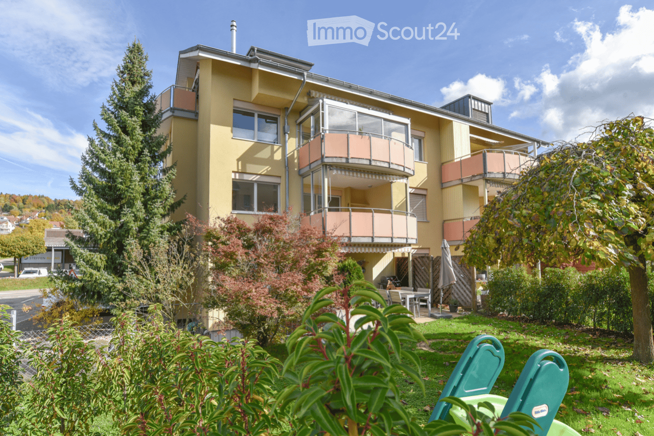 A three-story yellow building with multiple balconies and large windows, surrounded by greenery and a small patio area with outdoor furniture.