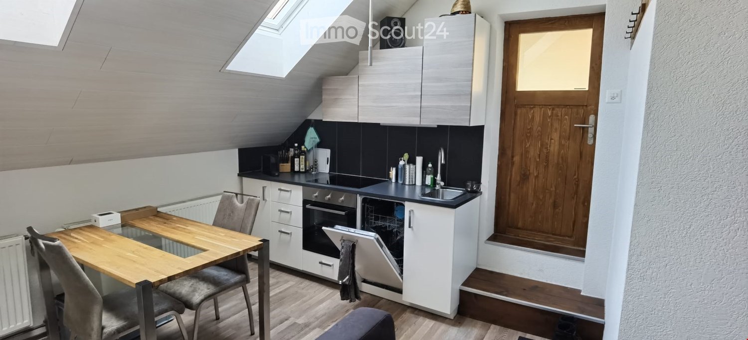Modern kitchen with white cabinets, wooden dining table, grey chairs, and built-in appliances including an oven and dishwasher. The space features a black countertop, a sink with a faucet, and a radiator under the window.
